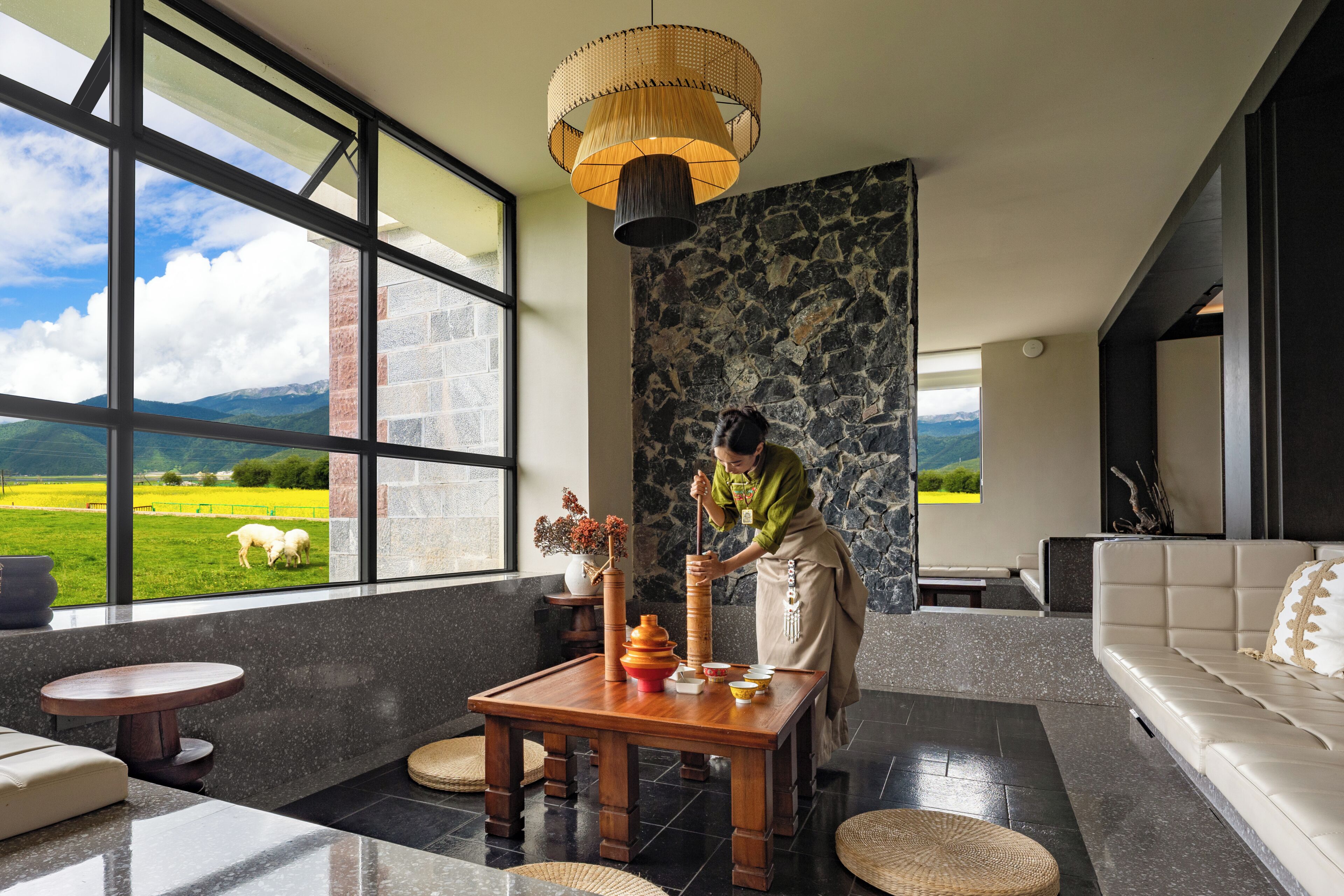 Un interior moderno con un ventanal grande, una pared de piedra, una mesa de madera y una persona preparando comida.