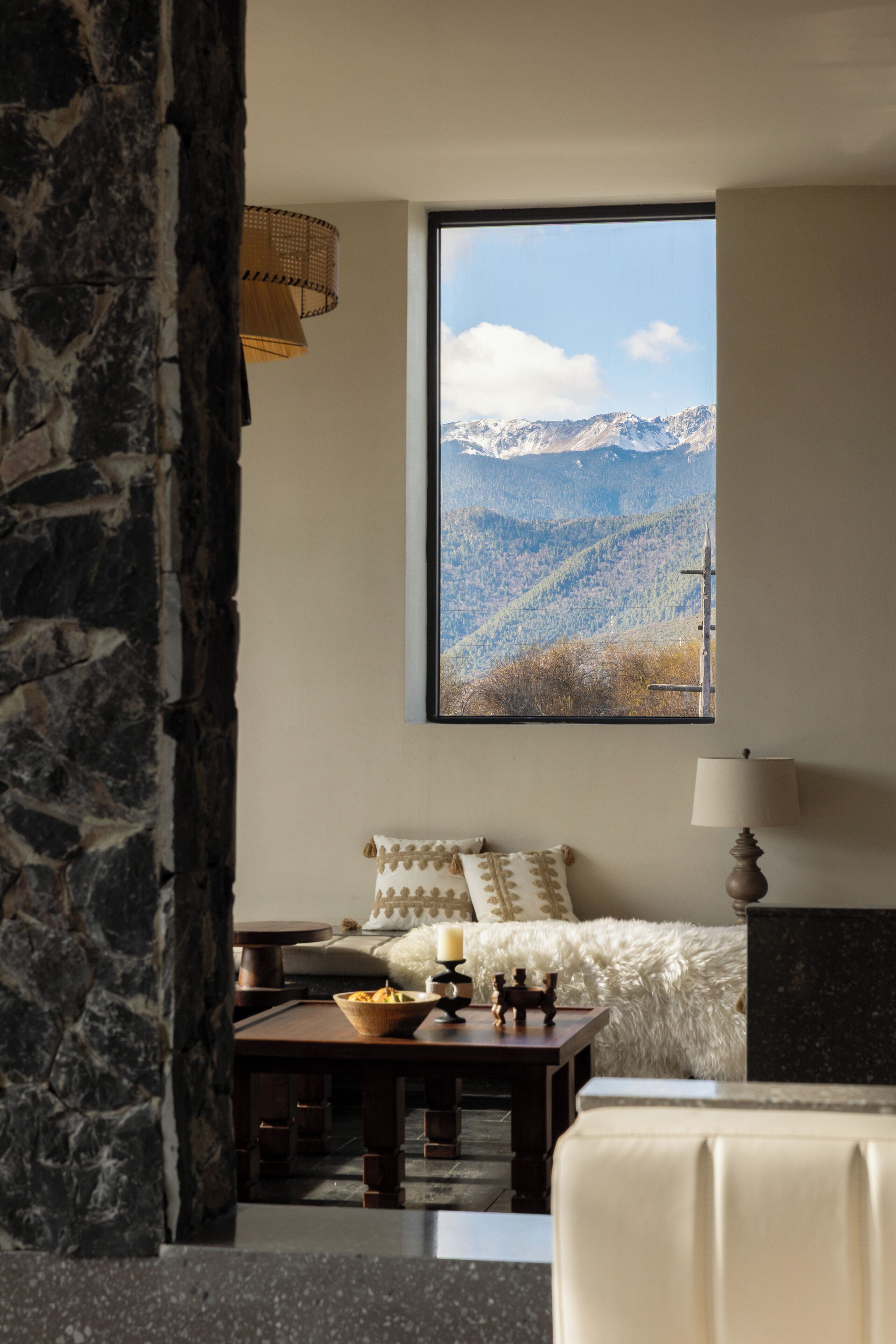 Ein Zimmer mit großem Fenster und Blick auf die Berge, einem Bett mit weißer Bettwäsche, einem Holztisch mit Schale und Kerzen sowie einer Steinstur.