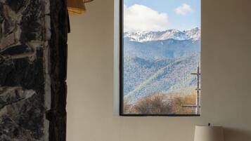 Ein Zimmer mit großem Fenster und Blick auf die Berge, einem Bett mit weißer Bettwäsche, einem Holztisch mit Schale und Kerzen sowie einer Steinstur.