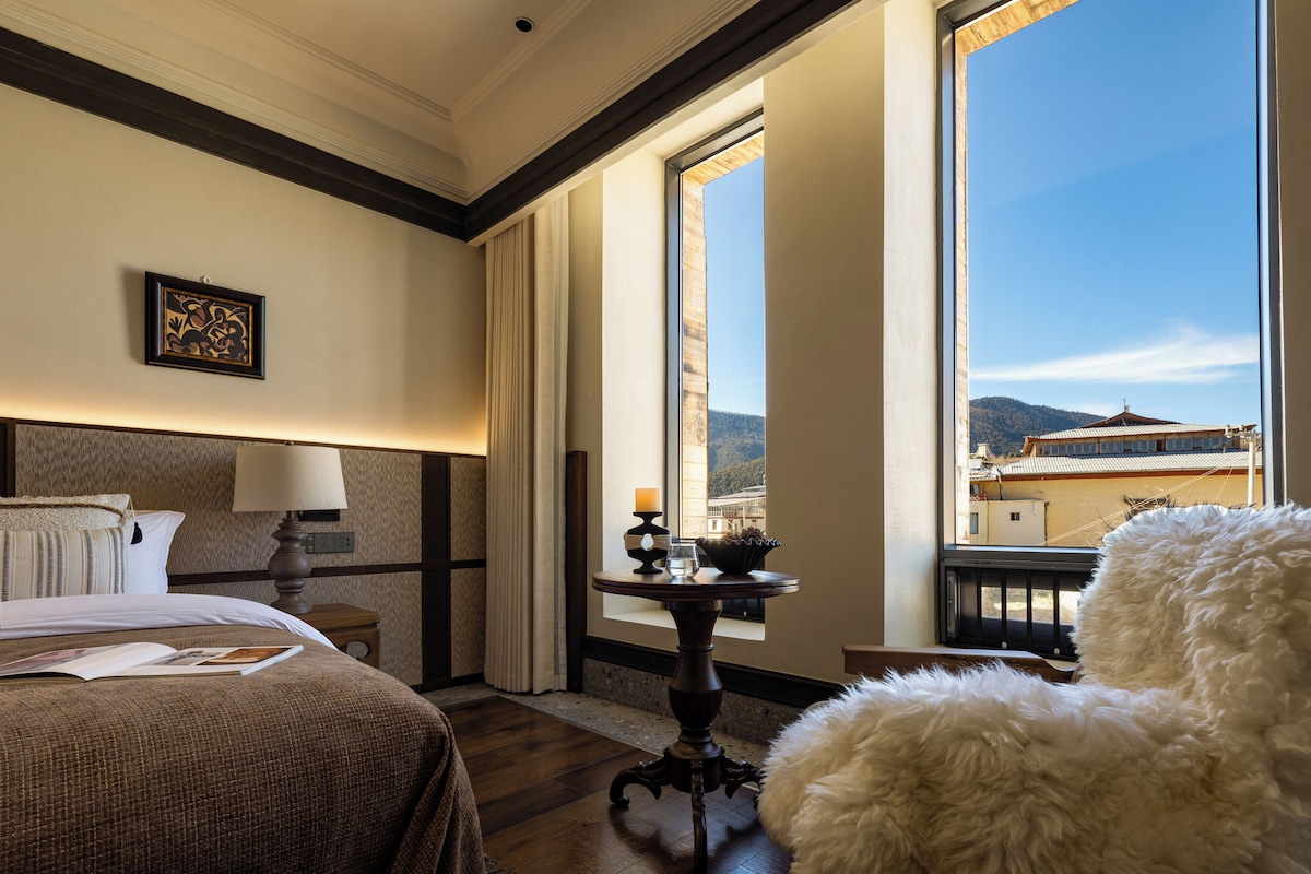 Una habitación de hotel con una cama, una mesita de noche con lámpara, un ventanal con vistas a edificios y montañas, y un sillón blanco mullido.