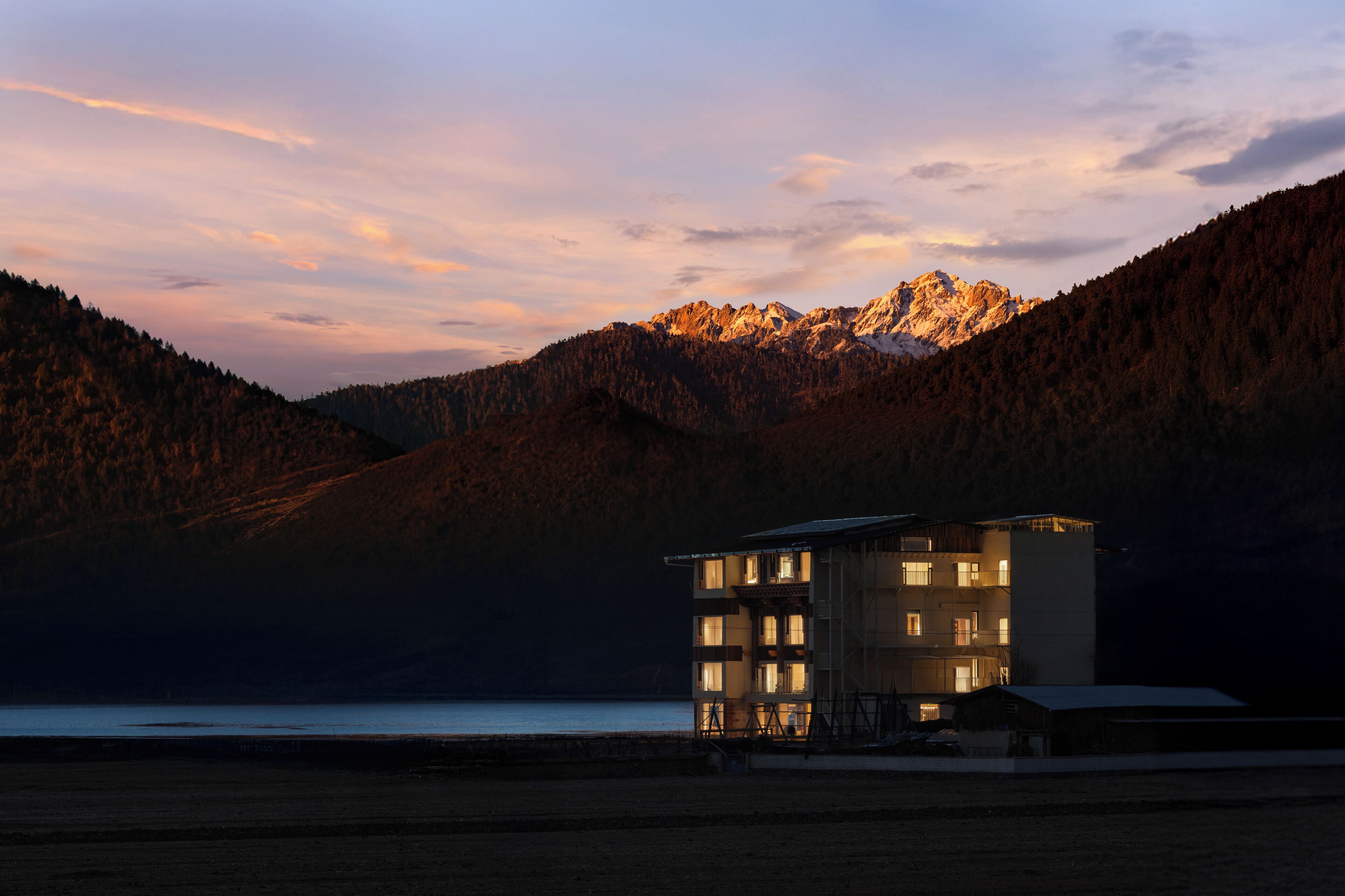 A building with multiple floors and balconies, situated near a body of water with mountains in the background.