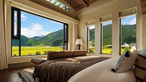 Quarto com amplas janelas, uma cama, mesa de cabeceira e vista para uma paisagem verde.