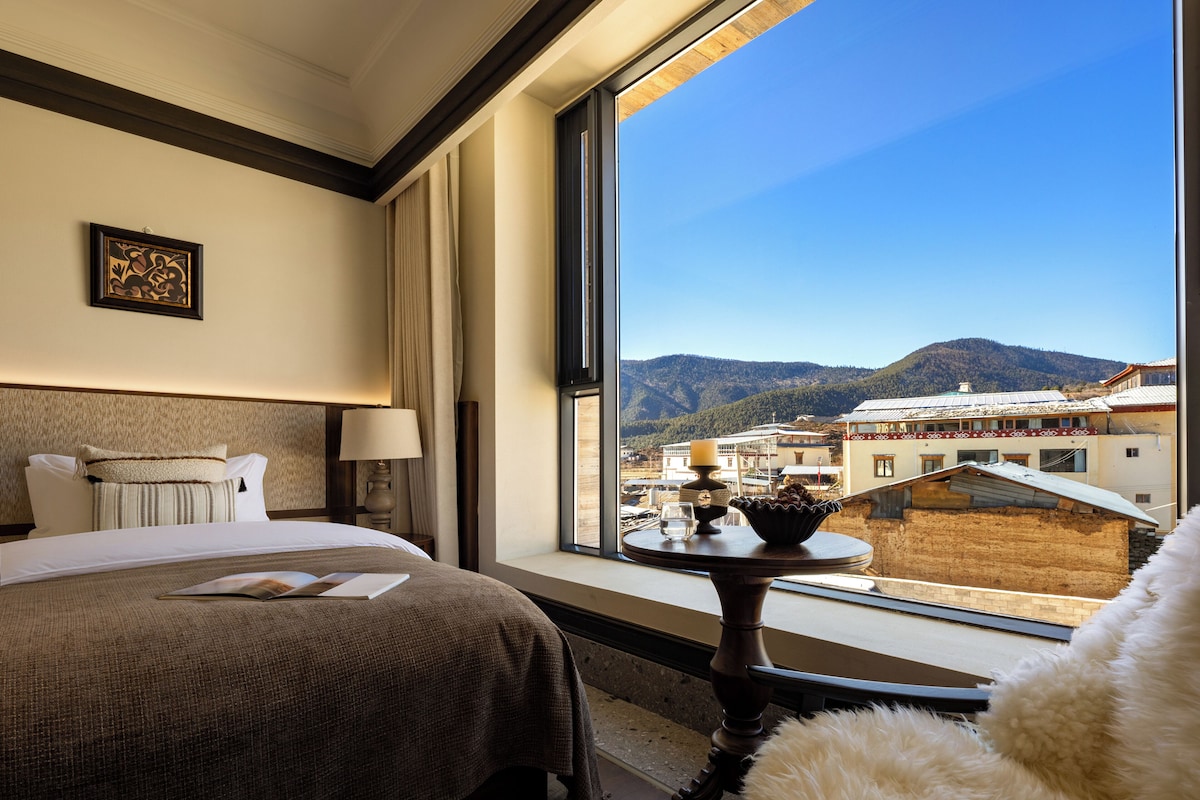 Habitación de hotel con un ventanal grande, una cama, una mesita de noche con lámpara y vistas a un paisaje montañoso.