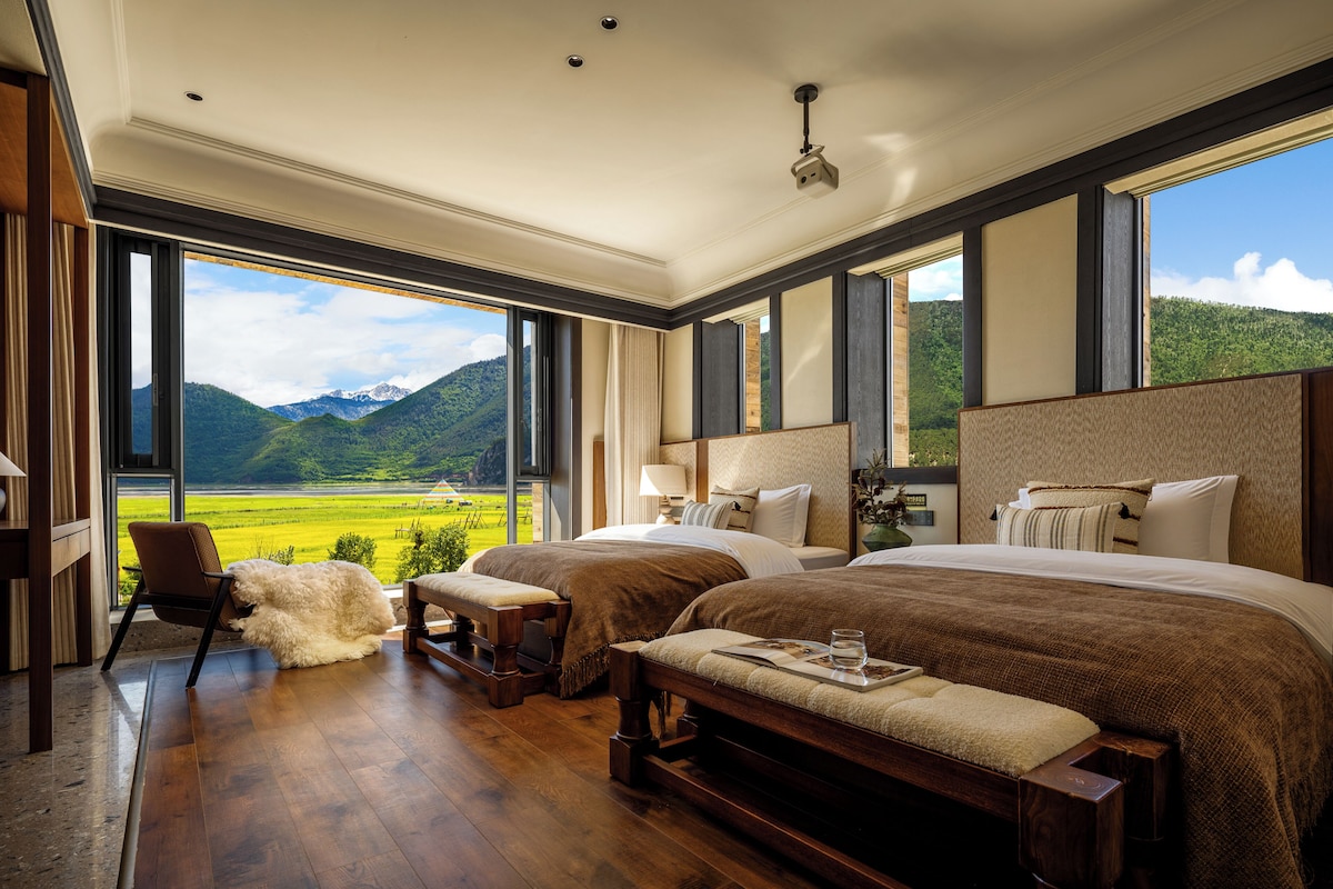 Un dormitorio amplio con una cama grande, una cama más pequeña, una silla y un banco. Se aprecia una vista de un paisaje verde a través de amplias ventanas.