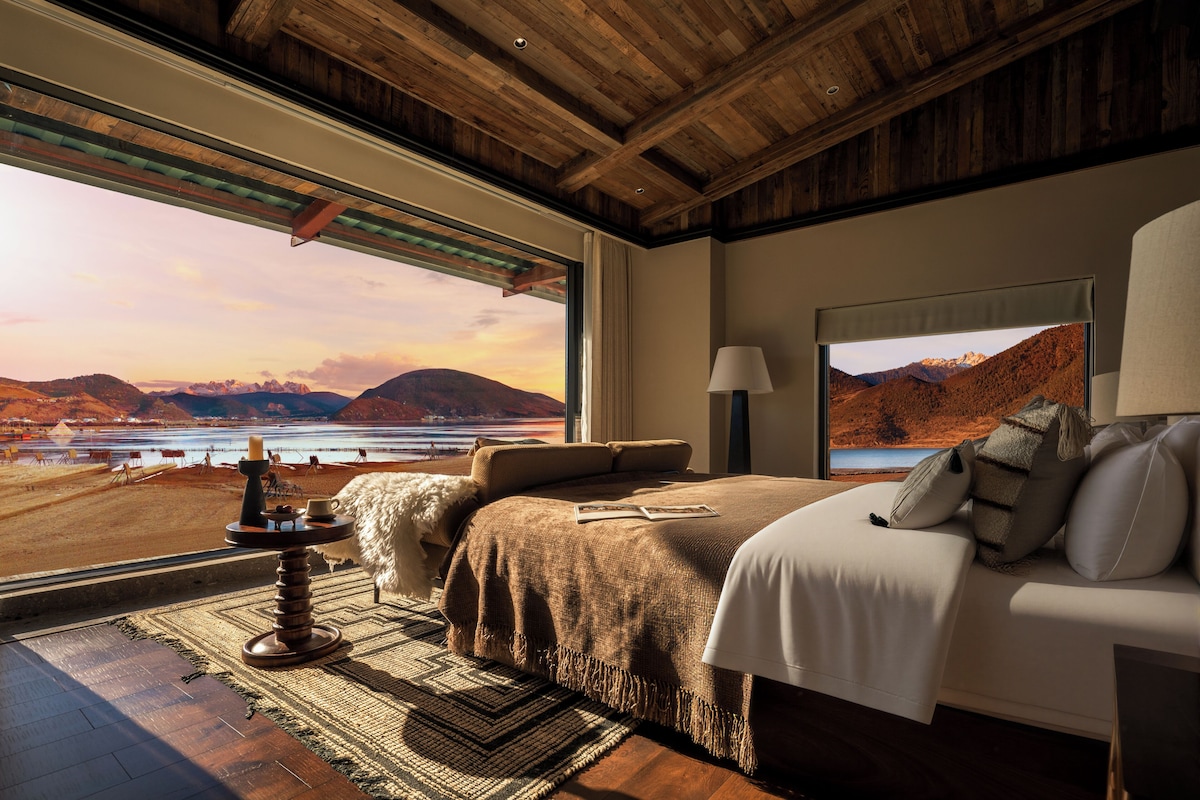 Un dormitorio con una ventana grande, una cama, una mesita de noche y vistas a montañas y un lago.