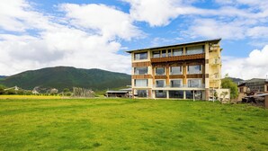 Um edifício de vários andares com grandes janelas e telhado plano, cercado por um campo gramado e montanhas ao fundo.