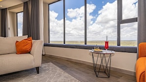 A room with a sofa, a small round table with a red vase, and a view of the sky through large windows. - Luxury and spacious 1BR Apartment at the Curve by the Park Nairobi Mombasa road. (Nairobi)