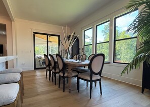 Zona pranzo con tavolo in legno, sei sedie e vista su una vegetazione attraverso ampie finestre.