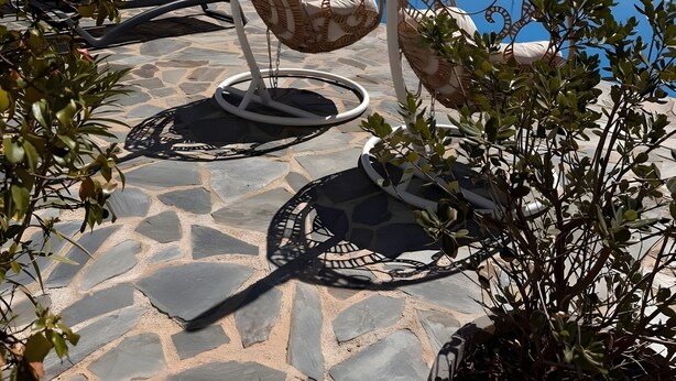 A poolside area with stone flooring, two wicker swing chairs, and a clear blue sky.