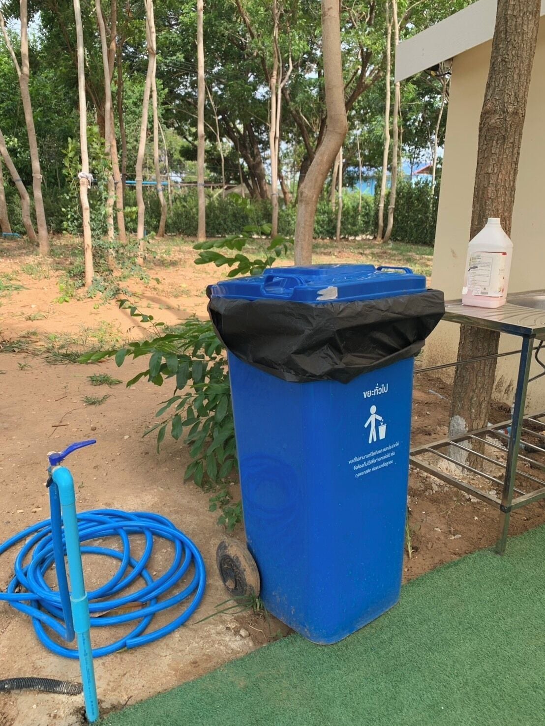 Ein blauer Mülleimer mit Recycling-Symbol, ein blauer Gartenschlauch und ein Tisch mit einem weißen Behälter.