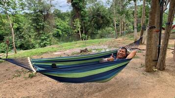 A hammock tied between trees in a natural setting with a dirt ground and a body of water in the background.