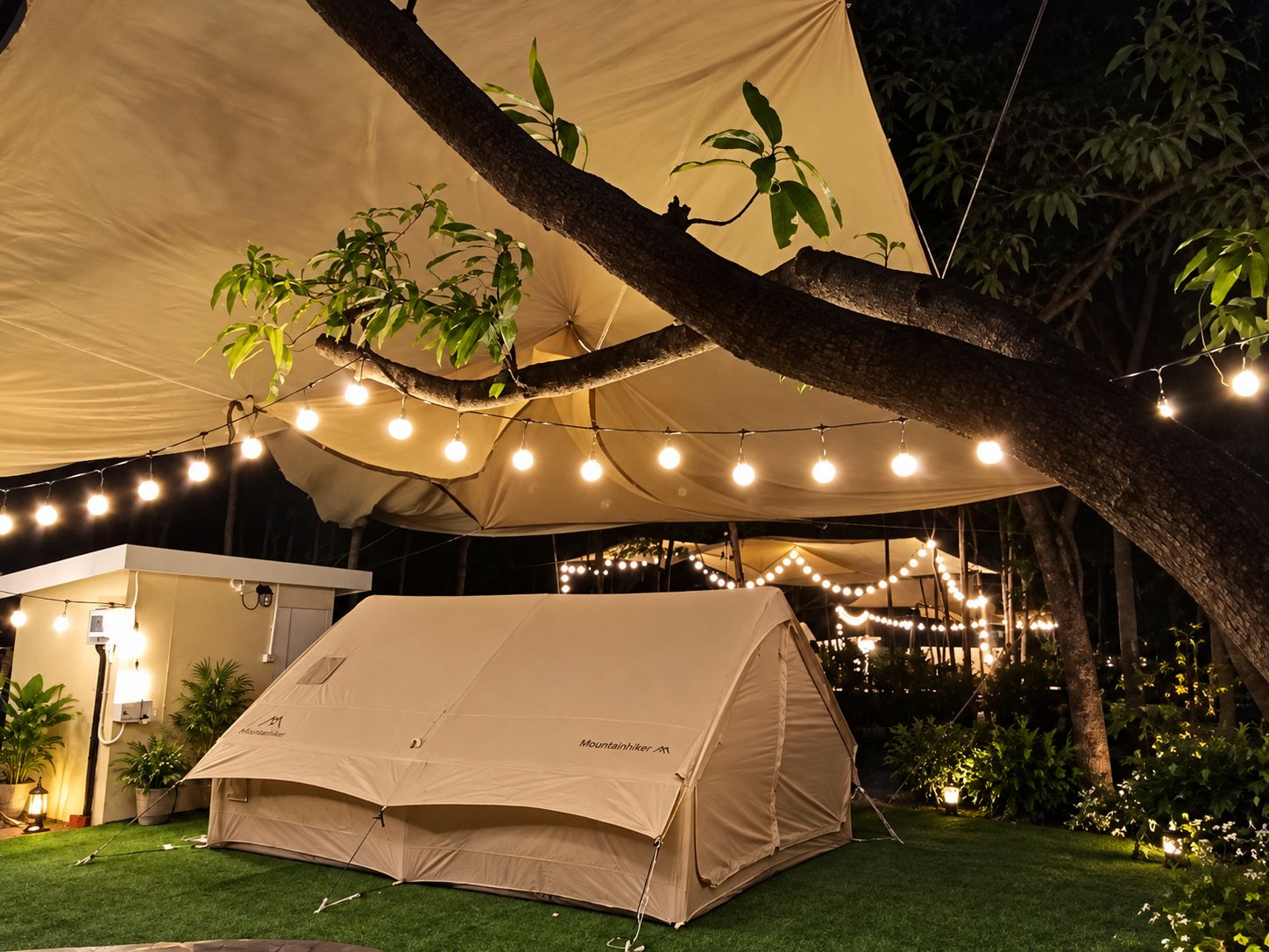A tent set up under a canopy with string lights, surrounded by greenery and a small building.