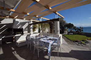 An outdoor patio with a table set for dining, a grill, and a view of the sea. - Villa Paola Caposuvero (Gizzeria Lido)