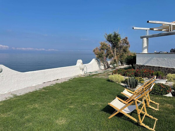 A grassy outdoor area with a white wall, a wooden bench, and a view of the sea.