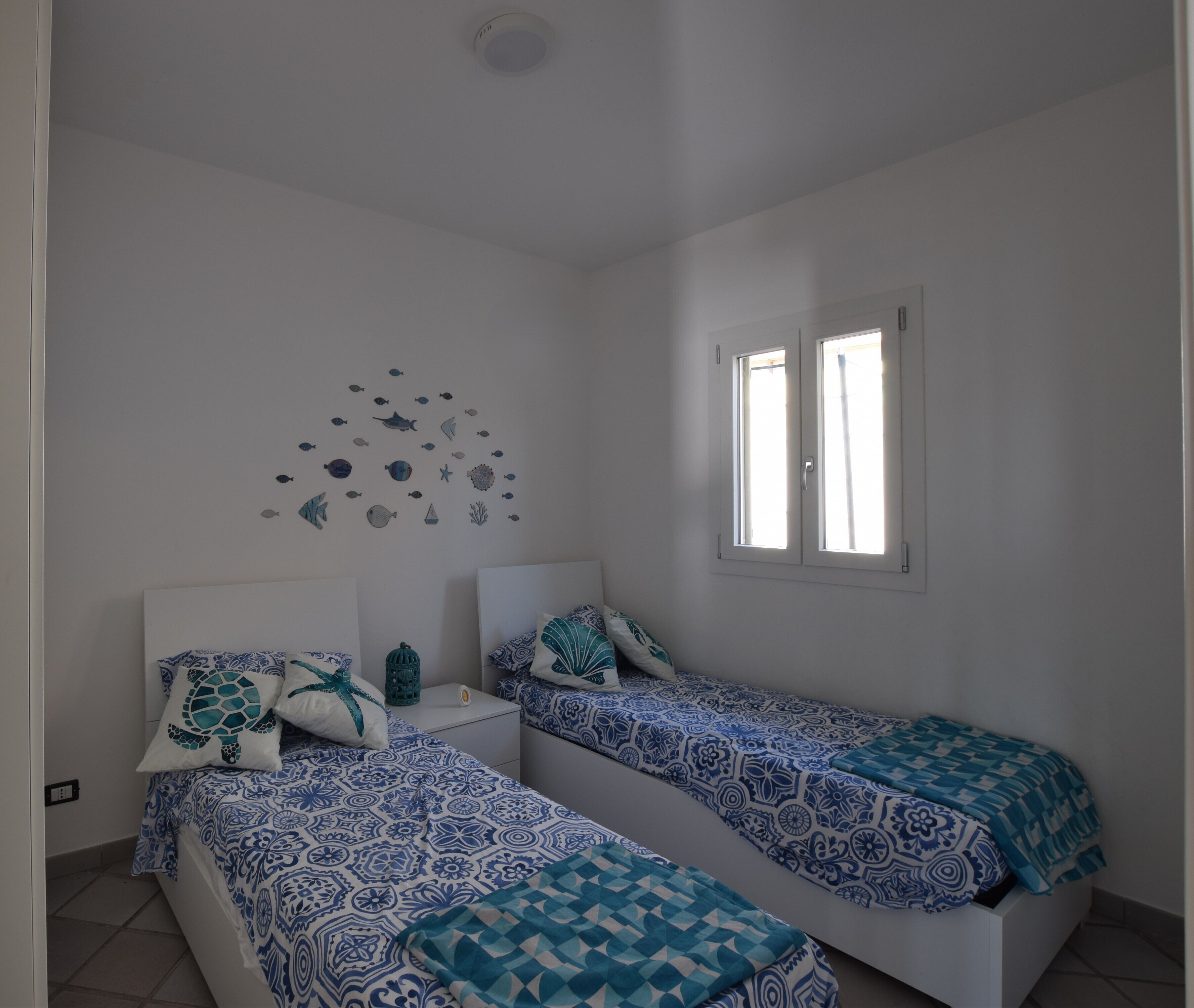 Une chambre avec deux lits, une décoration murale représentant des poissons et des créatures marines, et une fenêtre avec des stores.