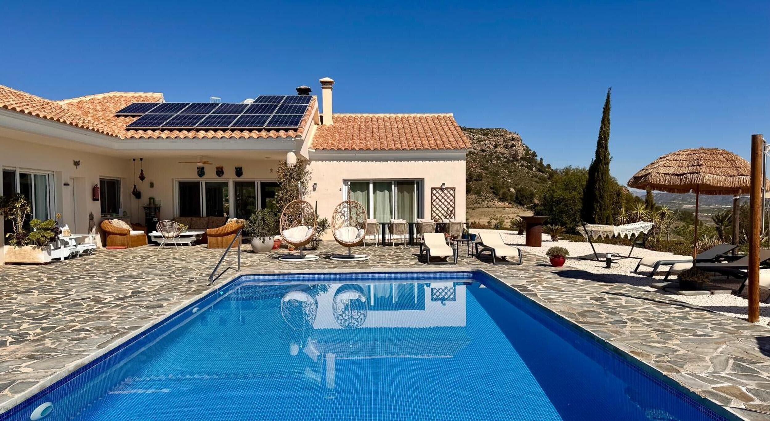 A pool area with a clear blue pool, lounge chairs, and a building with a tiled roof.