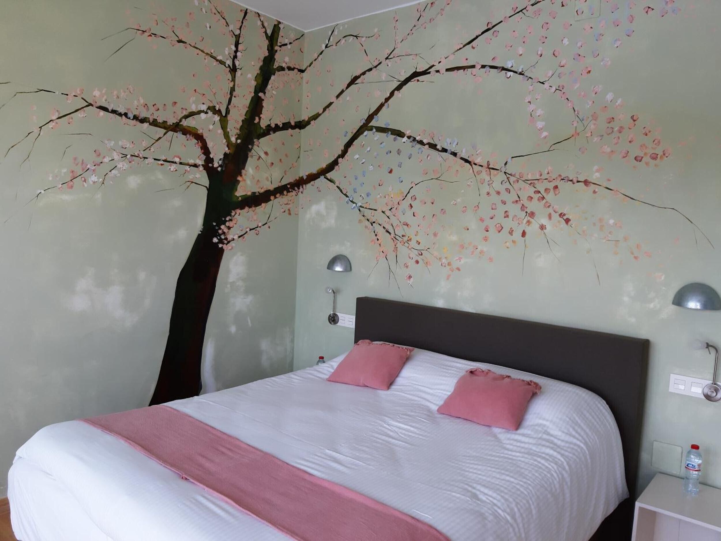 Un dormitorio con cama, almohadas, una mesita de noche, una lámpara de pared y una botella de agua sobre la mesita.