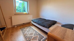 Une petite chambre avec un lit simple, une fenêtre, un radiateur, un tapis à motifs et une table en bois.
