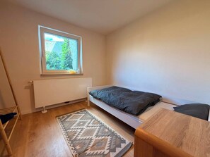 A small, single-bed room with a window, a radiator, a patterned rug, and a wooden table.