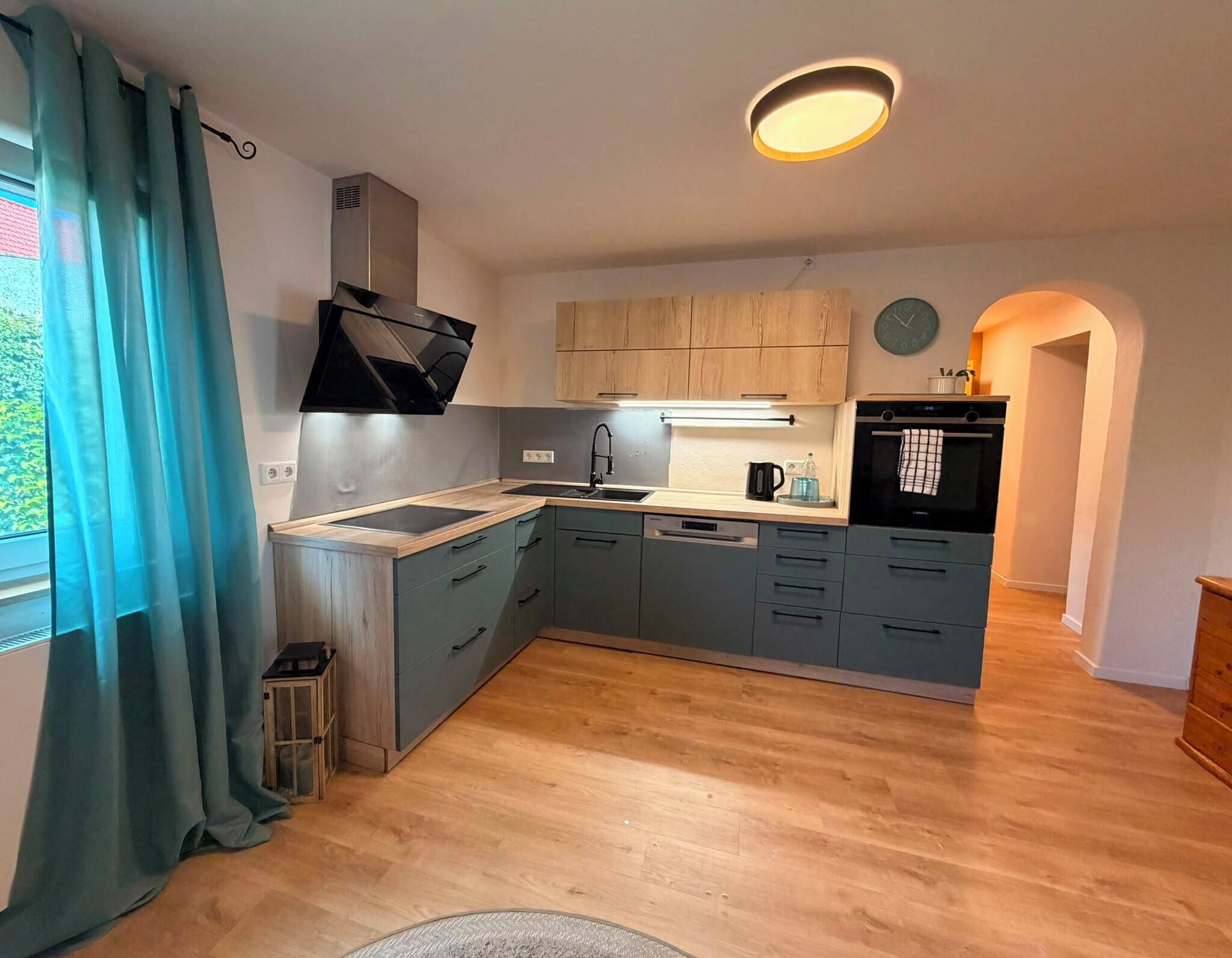 A modern kitchen with a built-in stove, a sink, and a dishwasher. There is a window with curtains, a clock on the wall, and a small trash bin.