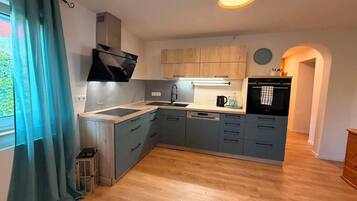 A modern kitchen with a built-in stove, a sink, and a dishwasher. There is a window with curtains, a clock on the wall, and a small trash bin.