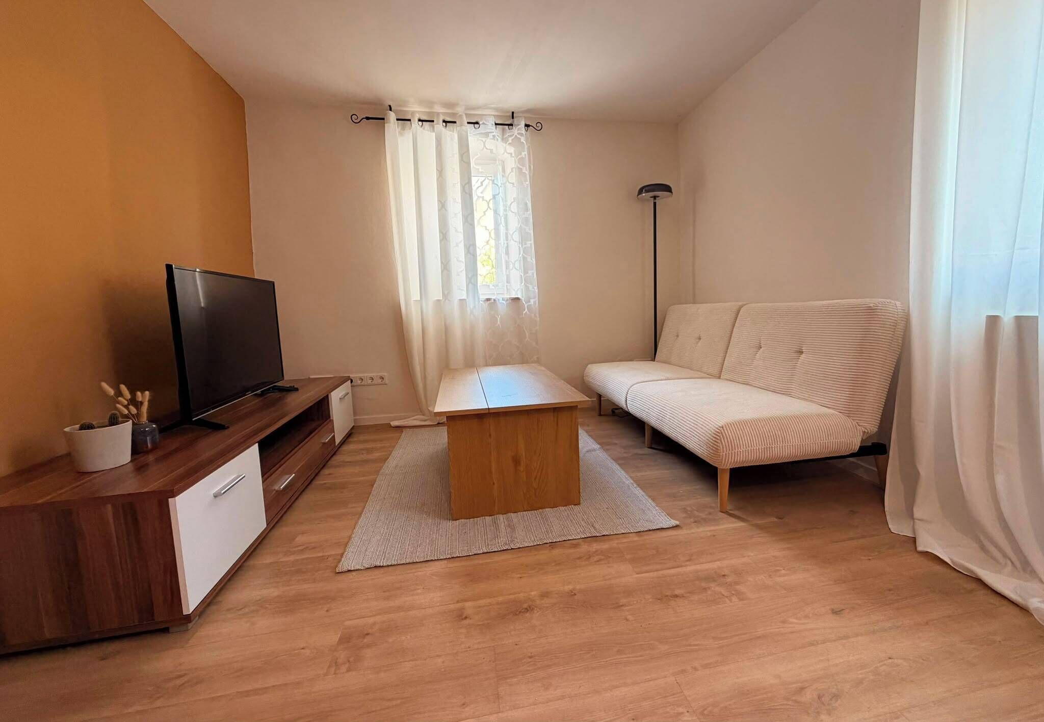 Un petit salon avec un téléviseur posé sur un meuble en bois, un canapé, une table basse et une fenêtre avec des rideaux.