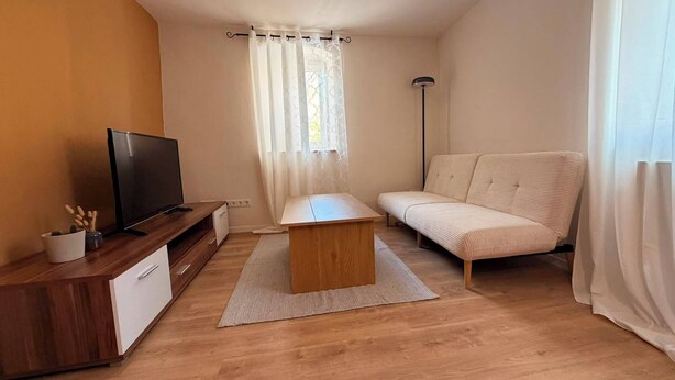 Un petit salon avec un téléviseur posé sur un meuble en bois, un canapé, une table basse et une fenêtre avec des rideaux.