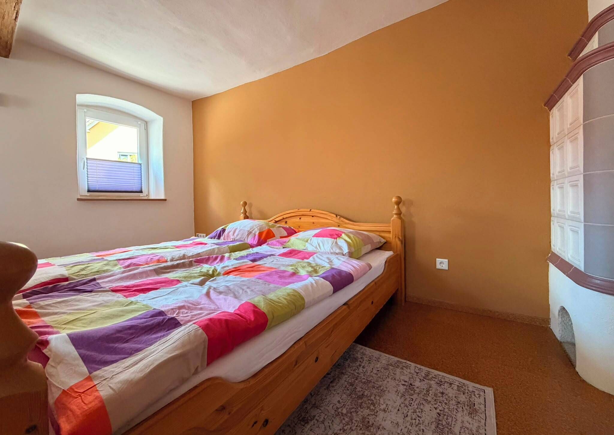 Une chambre avec un lit en bois, une courtepointe aux couleurs vives, une petite fenêtre et un mur blanc.