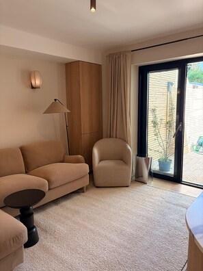 A living room with a sofa, armchair, and a small table. There is a lamp, a cabinet, and a sliding glass door leading to an outdoor area. - Hackney Garden Apartment (London)