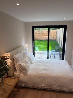 A bedroom with a bed, a bedside table with a lamp, a potted plant, and a view of a garden through a sliding door. - Hackney Garden Apartment (London)