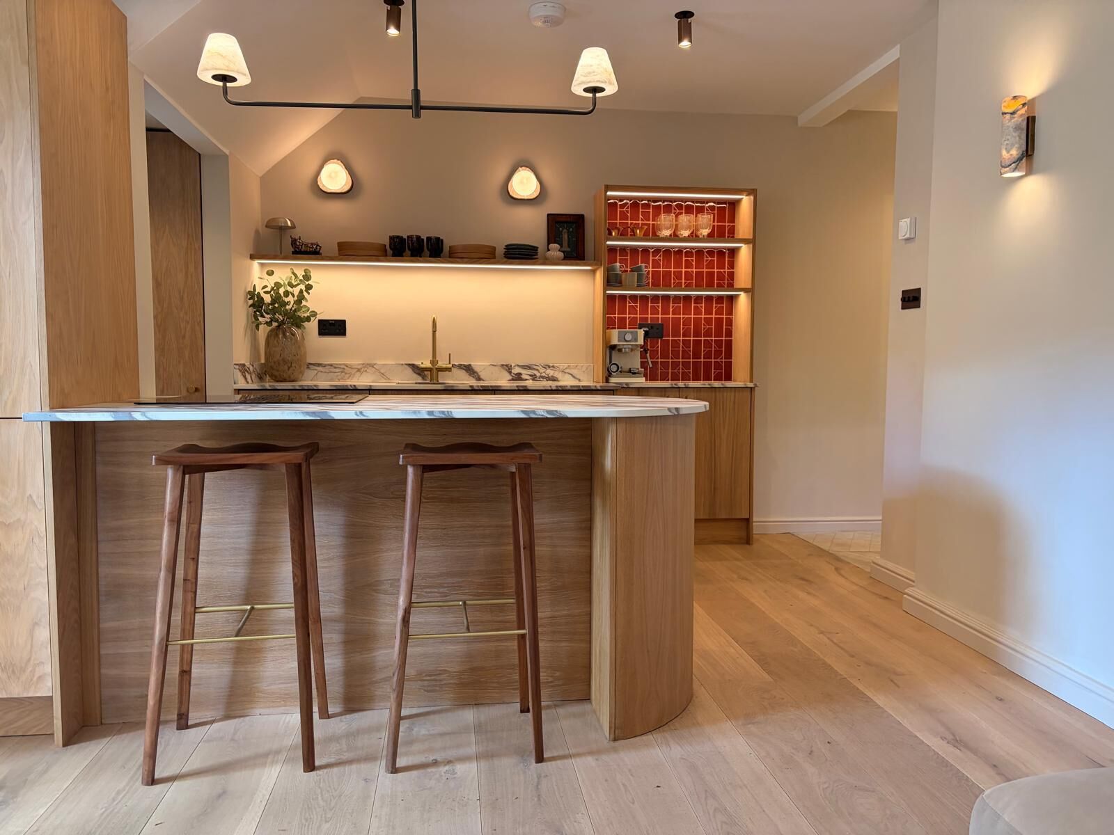 Une cuisine moderne dotée d’un îlot central, de tabourets en bois, de luminaires suspendus et d’un pare-feu rouge.