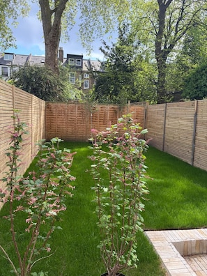 A backyard with a wooden fence, green grass, and flowering plants. - Hackney Garden Apartment (London)