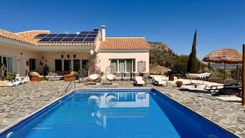 A pool area with a clear blue pool, lounge chairs, and a building with a tiled roof.