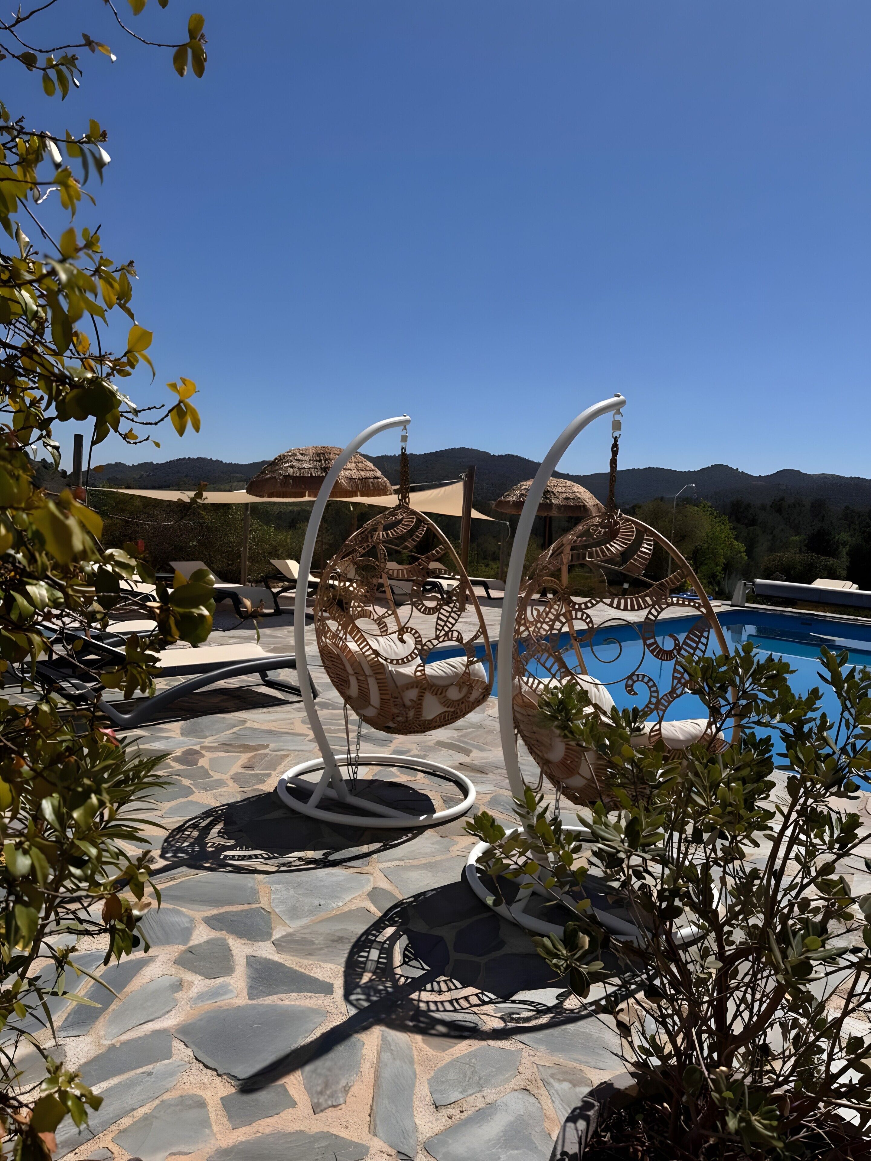 Zona junto a la piscina con suelo de piedra, dos sillones de mimbre y un cielo azul despejado.