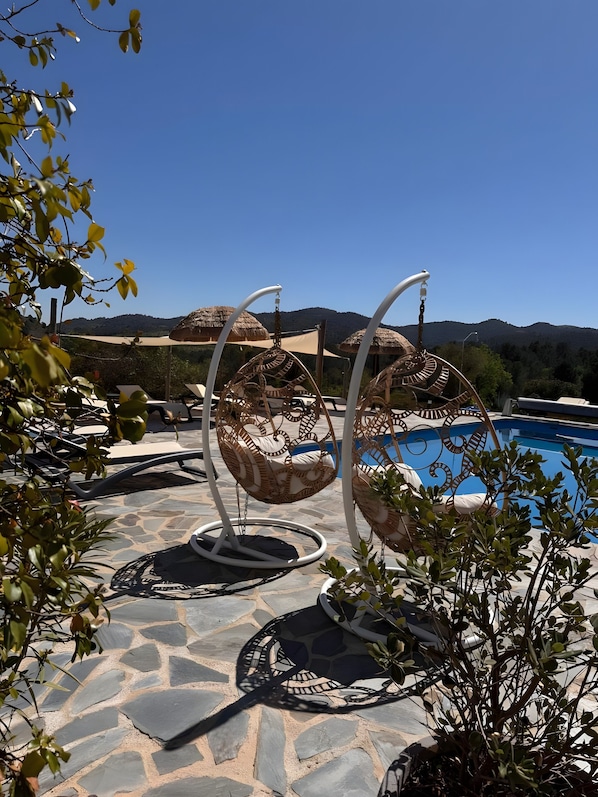 Zona junto a la piscina con suelo de piedra, dos sillones de mimbre y un cielo azul despejado.