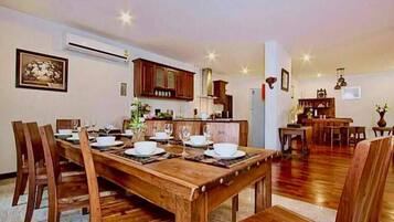 A dining area with a wooden table set for four, flanked by wooden chairs, a kitchen in the background, and a living area with a sofa and coffee table.