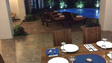 Un coin repas avec vue sur la piscine, comprenant une table dressée pour deux personnes, avec assiettes, couverts et verres.