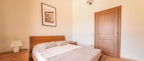 A bedroom with a wooden bed, a nightstand, a lamp, a framed picture on the wall, and a wooden door.