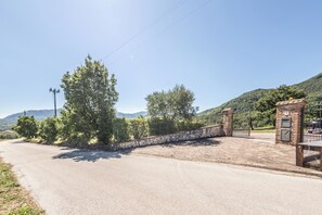 Une route goudronnée mène à une entrée fermée par un portail, elle-même bordée d’un mur en pierre et surmontée d’un portail en métal.