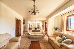 A spacious living room with a chandelier, leather sofas, a wooden coffee table, and a large window with curtains.