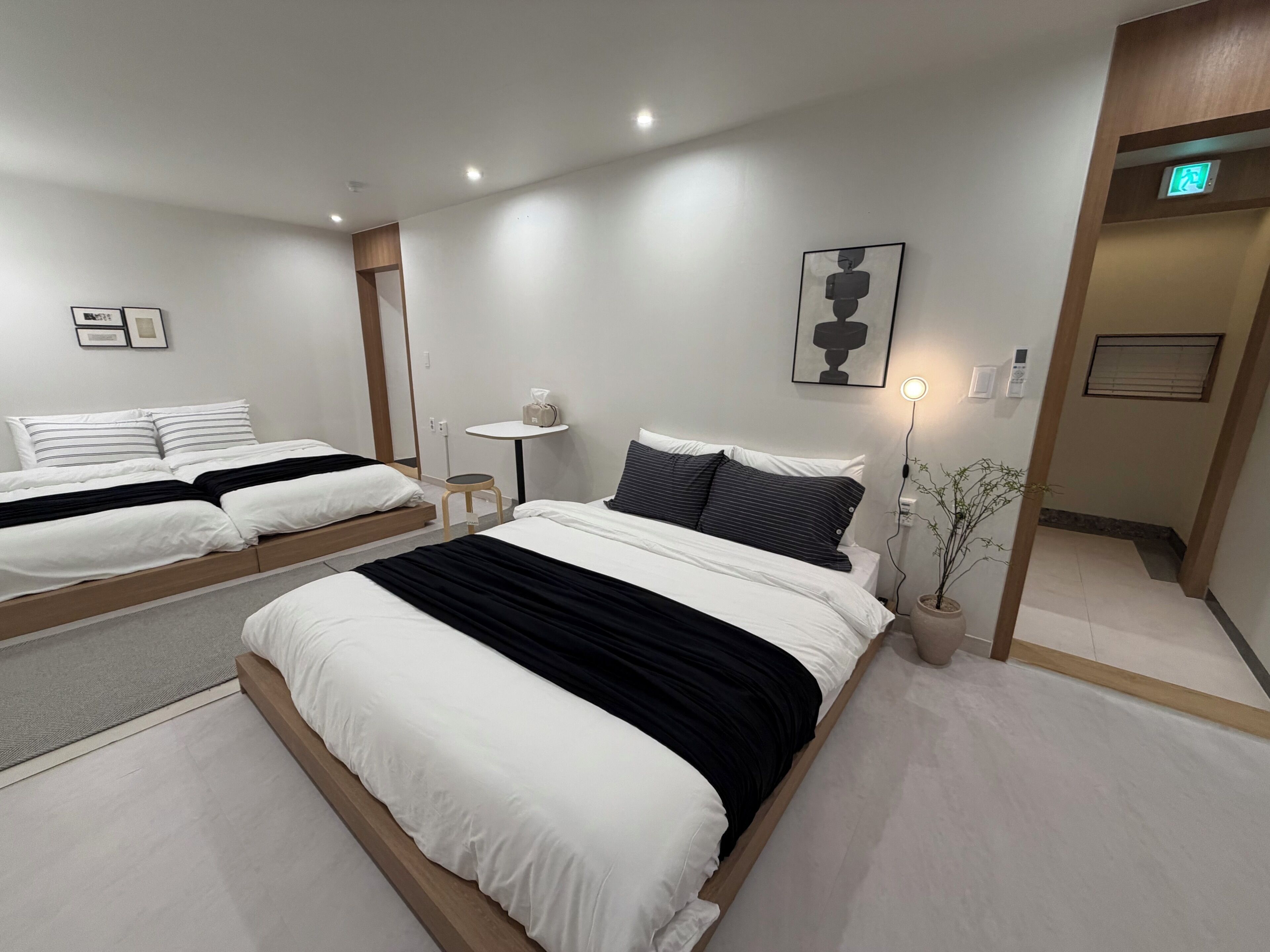 A modern hotel room with two beds, a small table, a wall-mounted light, and a potted plant.
