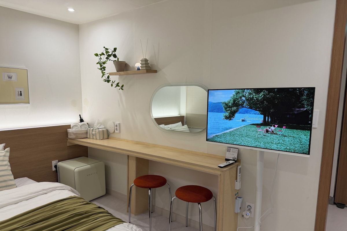Een hotelkamer met een bed, een bureau met twee krukken, een televisie, een spiegel en een kleine potplant.