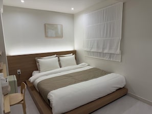Een hotelkamer met een bed, een houten hoofdeinde, een nachtkastje met een stoel en een raam met jaloezieën.