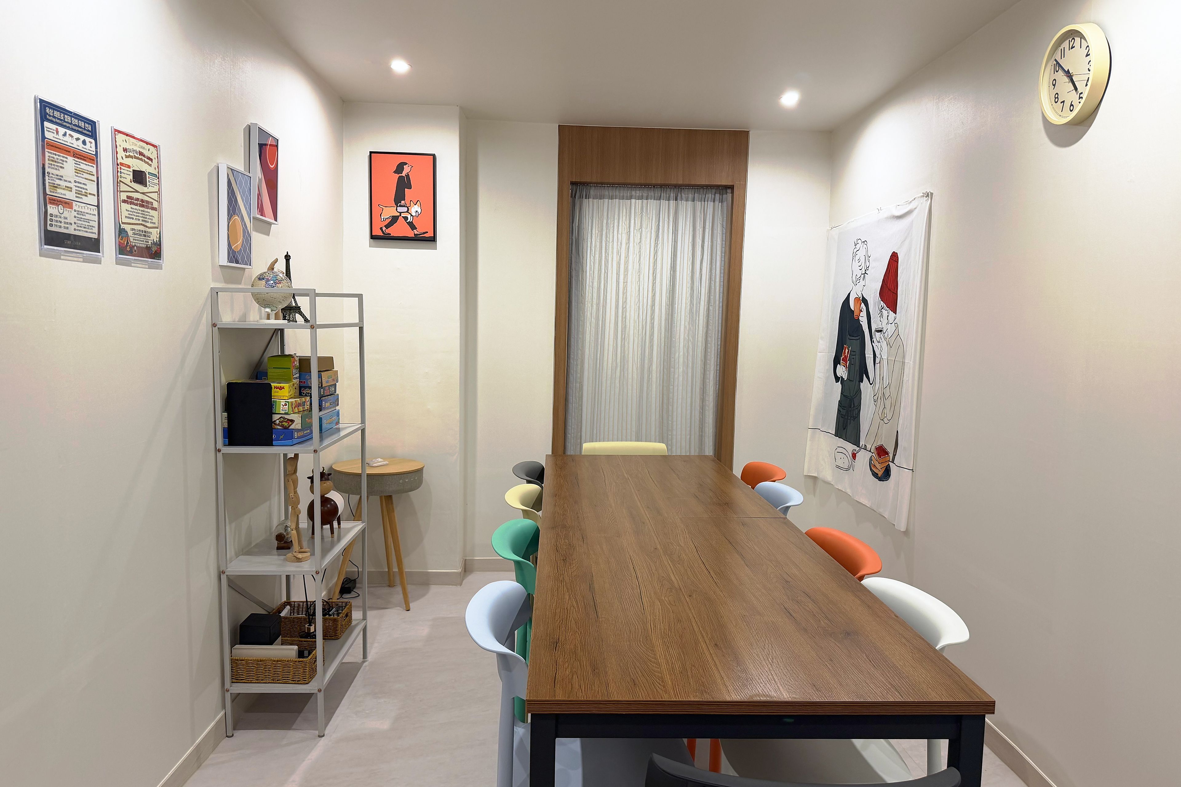 A dining area with a wooden table, colorful chairs, a shelf with boxes, and a clock on the wall.