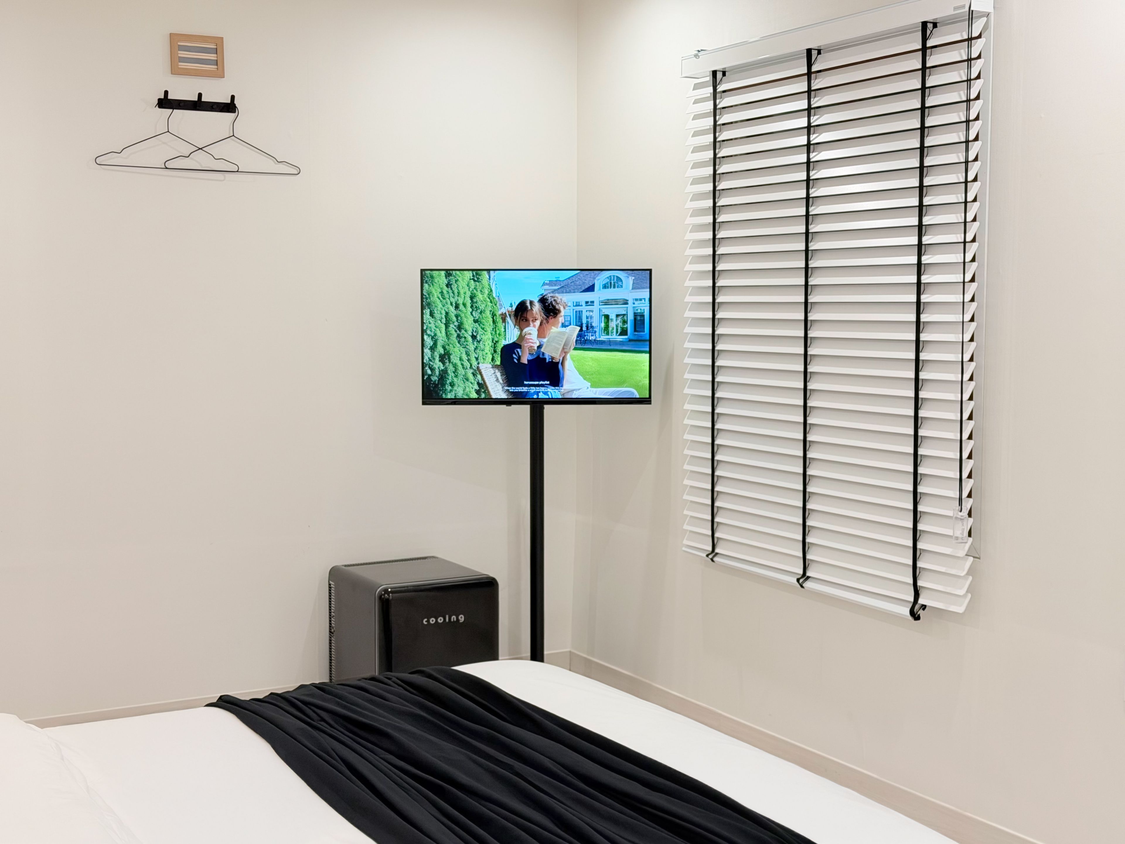 A hotel room with a bed, a television mounted on the wall, a window with blinds, and a small speaker.
