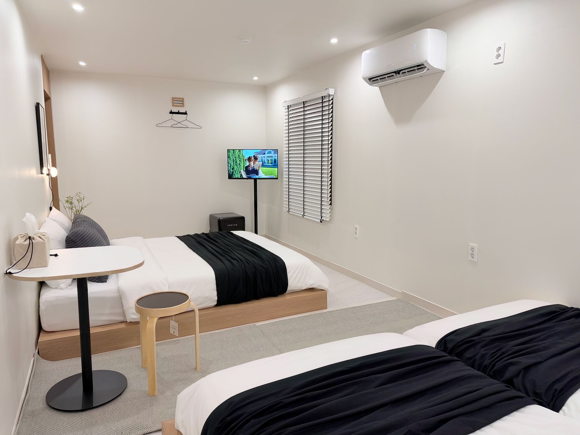 A hotel room with a bed, bedside table, lamp, TV, air conditioning unit, and a window with blinds.