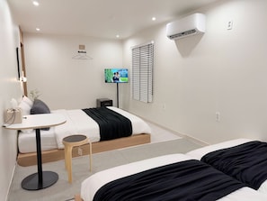 Habitación de hotel con cama, mesita de noche, lámpara, televisor, equipo de aire acondicionado y ventana con persianas.