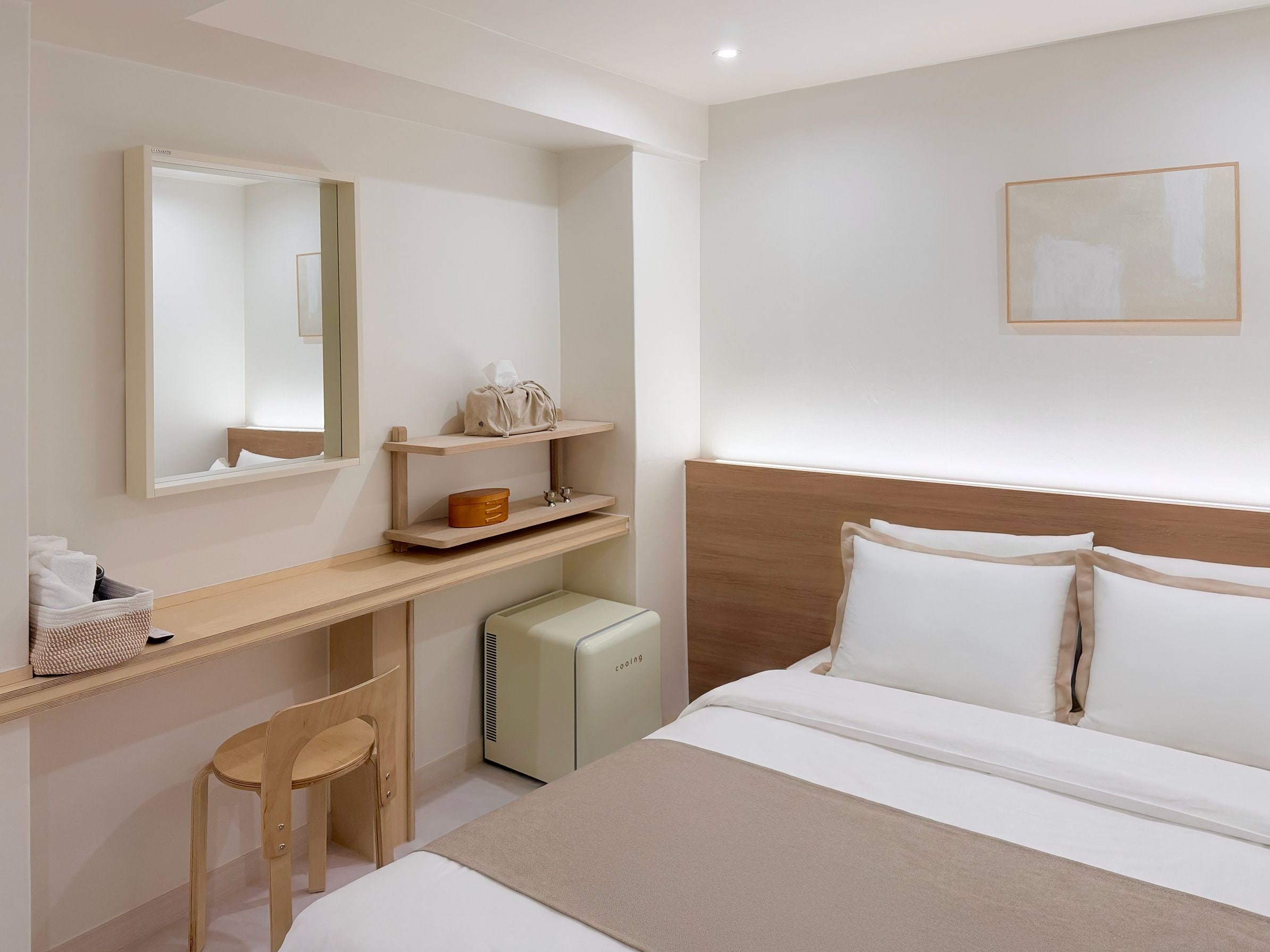 Habitación de hotel con cama, escritorio, silla, mesita de noche, espejo, pequeño calentador y un cuadro en la pared.