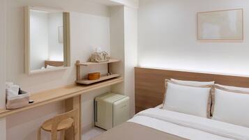 A hotel room with a bed, a desk, a chair, a bedside table, a mirror, a small heater, and a framed picture on the wall.