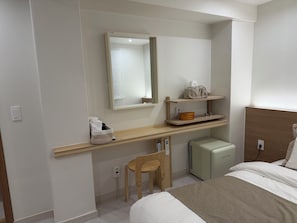 Een hotelkamer met een bed, een klein bureau met een stoel, een spiegel, een plank met toiletartikelen en een kleine airconditioning.